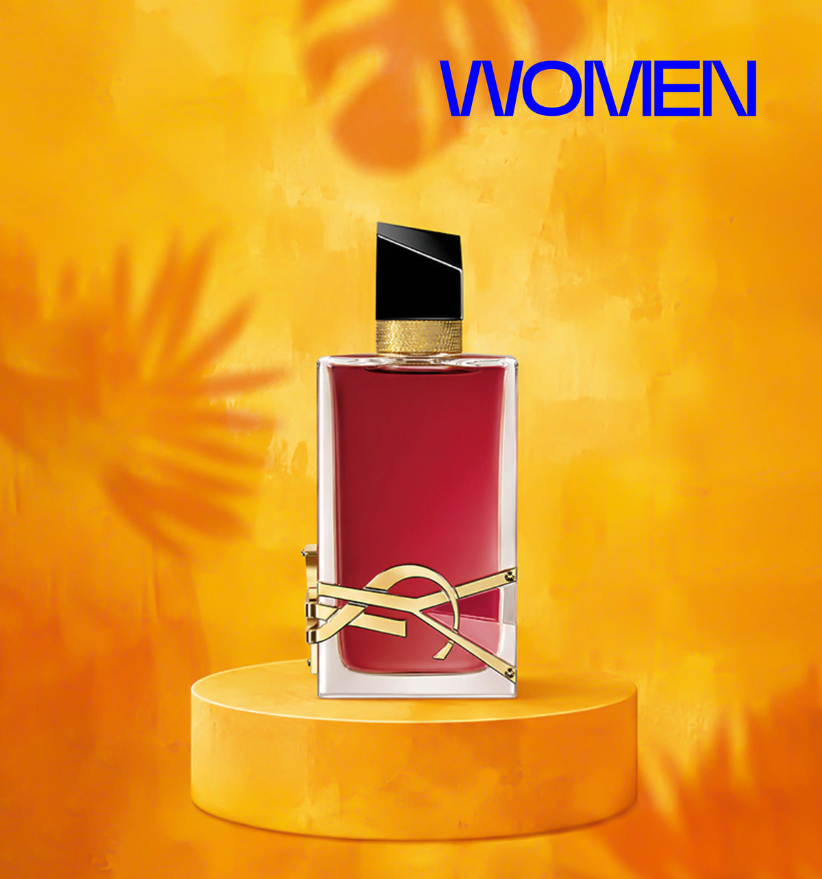A rectangular perfume bottle with a deep pink-red liquid, a black cap, and a gold YSL logo, displayed on a matching yellow pedestal against a warm yellow-orange background with the word 'WOMEN' in blue at the top.